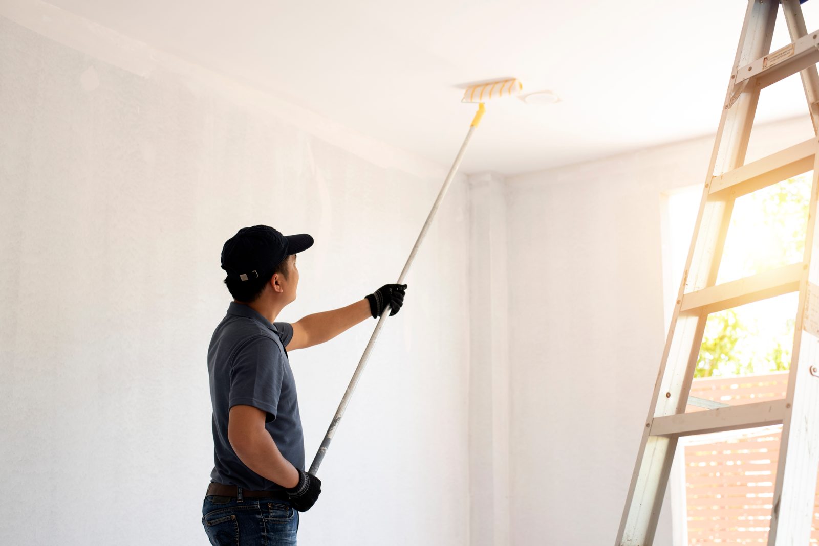Professional painter rolling paint onto an interior wall and ceiling beside a ladder